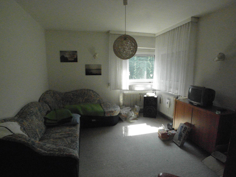 Ein Wohnzimmer mit einer Couch und einem Fernseher; auf dem Boden liegt viel Kleinkram